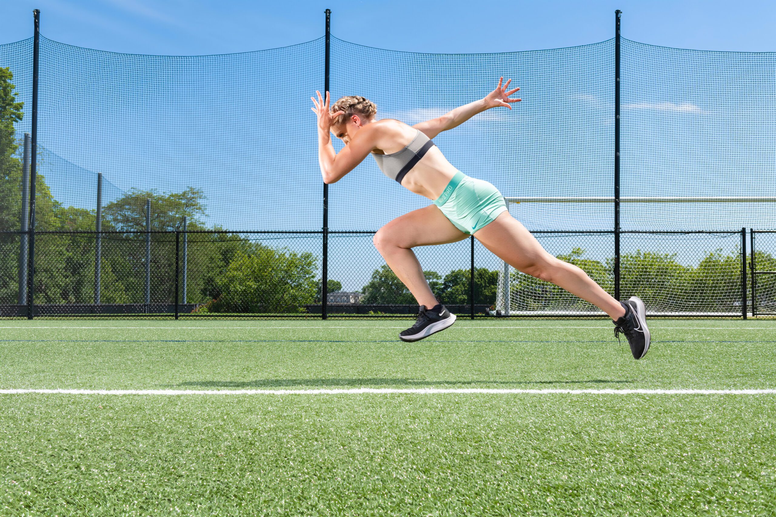 A person in athletic wear sprints across a grassy sports field on a sunny day. The runner is in a high-motion pose, with one arm forward and the other back. A black fence and trees are in the background under a clear blue sky.
