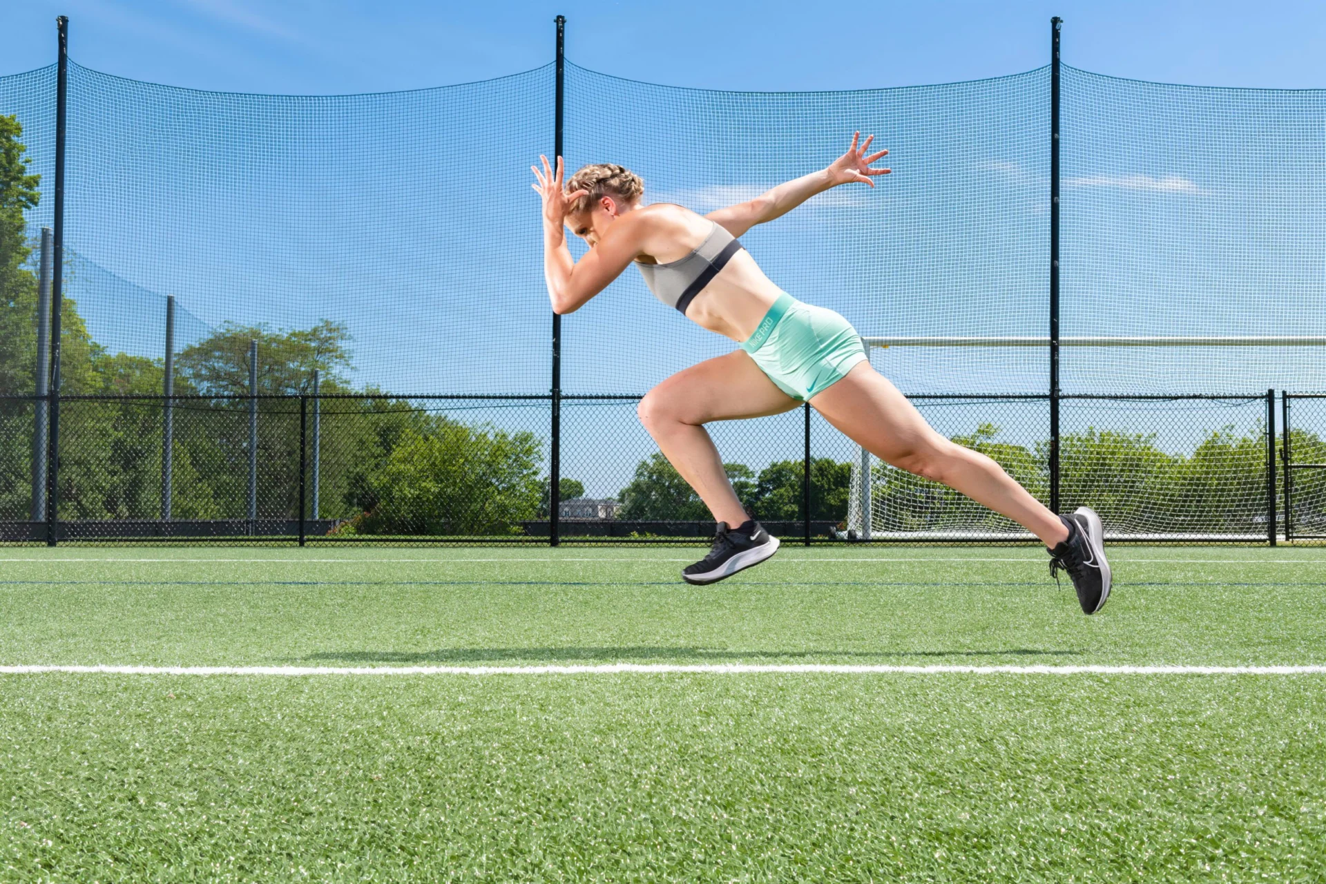 A person in athletic wear sprints across a grassy sports field on a sunny day. The runner is in a high-motion pose, with one arm forward and the other back. A black fence and trees are in the background under a clear blue sky.