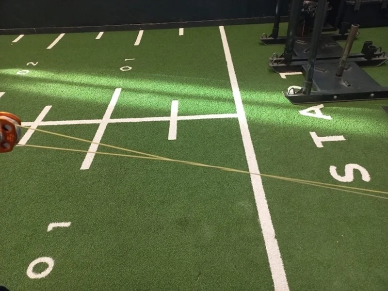Green turf with white yard lines resembling a football field, gym equipment on the right, and a resistance band being pulled diagonally across the image from left to right.