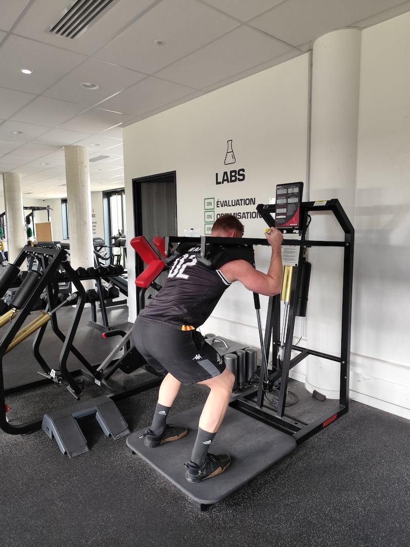 A person in sportswear is using a squat machine in a gym. They are performing a squat exercise with their back against a padded support while holding onto handles. The room is well-lit with gym equipment in the background.