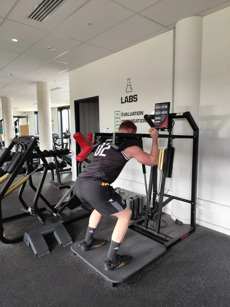 A person in sportswear is using a squat machine in a gym. They are performing a squat exercise with their back against a padded support while holding onto handles. The room is well-lit with gym equipment in the background.