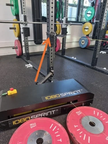 A gym squat rack with various colored weight plates is shown. An orange arrow points to a mechanism on the rack. In front, a black 1080 Sprint machine and pink 25 kg weight plates are visible on the floor.