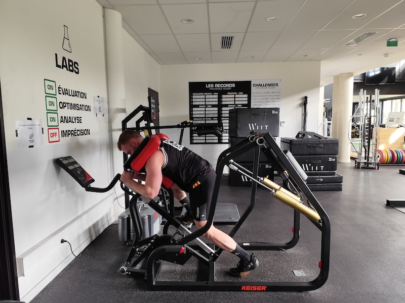 A man in athletic gear uses a Keiser exercise machine in a gym. Hes in a leaning position, pushing against a padded bar. The wall has signs labeled Labs and a motivational board, and there are weights and equipment in the background.