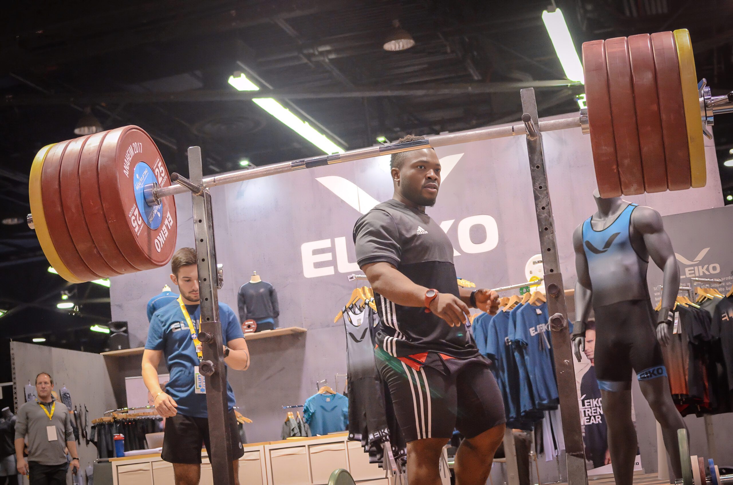 A weightlifter prepares to lift a heavily loaded barbell during a competition. A person stands nearby monitoring. Various gym apparel and equipment are displayed in the background.