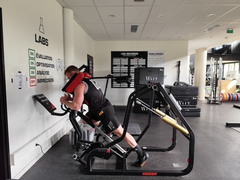 A person is using a leg press machine in a gym. They are wearing a black tank top and shorts. The gym has various exercise equipment, informational posters on the wall, and a rubberized floor.
