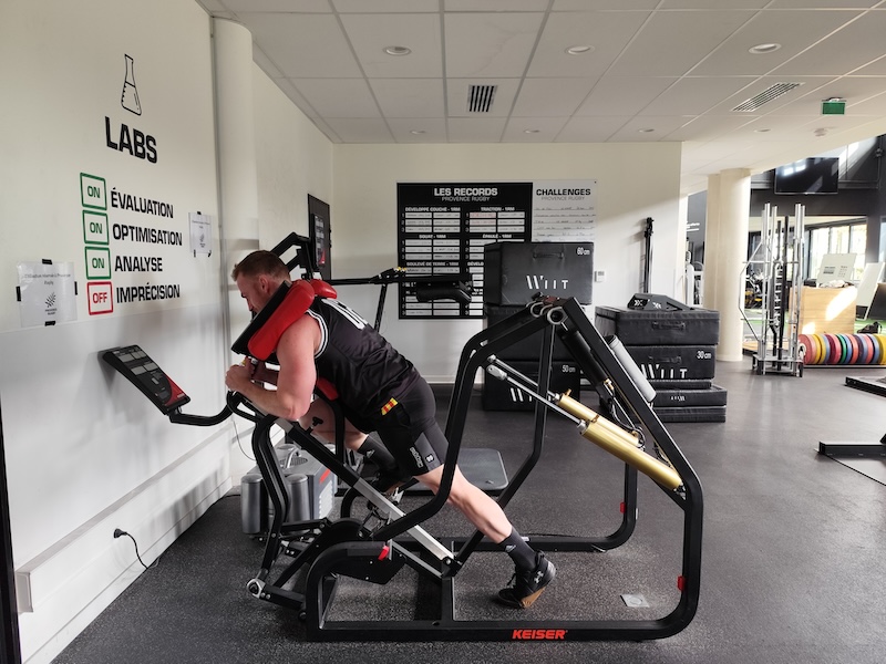 A person exercises on a Keiser machine in a gym. The room has records displayed on one wall and various equipment like weights and boxes scattered around. The gym is well-lit, with a clean, modern interior design.