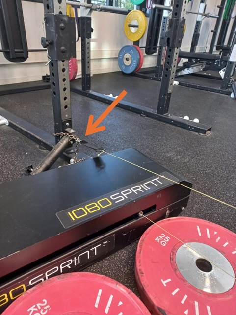 A 1080 Sprint training device is set up in a gym with red weight plates nearby. An orange arrow points to a metal chain attachment connecting the device to a squat rack.