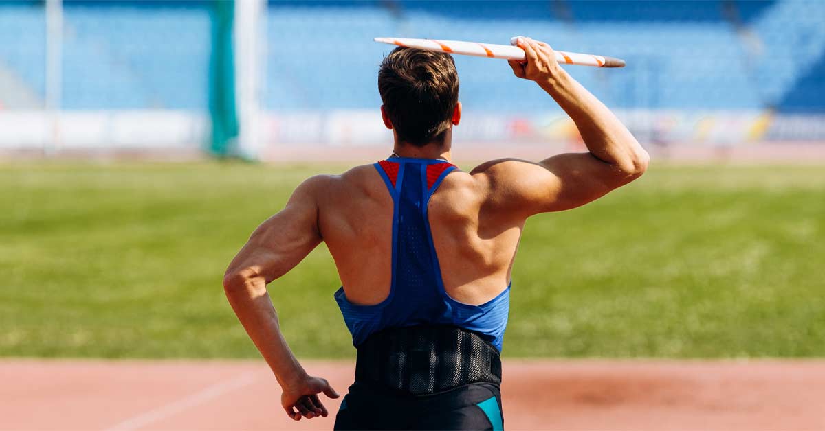Athlete in a blue tank top preparing to throw a javelin at a stadium. The person is seen from behind