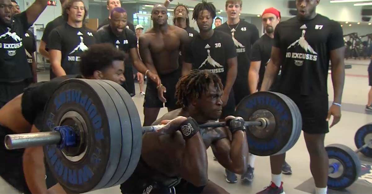 Max Out Drop|Johnny Parker|Russian Lifts|General Adaptation Syndrom|A person in a gym is performing a heavy squat with a barbell loaded with several weight plates. They are wearing a black T-shirt and red shoes