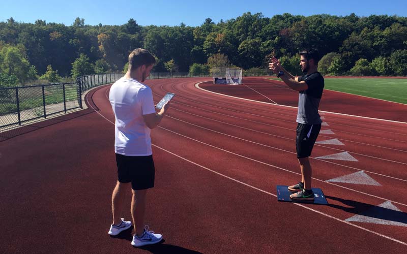 Jump Mat|Raptor Test using GymAware|A man in athletic clothing performs an exercise on a green turf in front of a red wall with EXCEED Sports Performance & Fitness written on it. Orange cones are lined up on the right.|A person stands on a mat in a gym