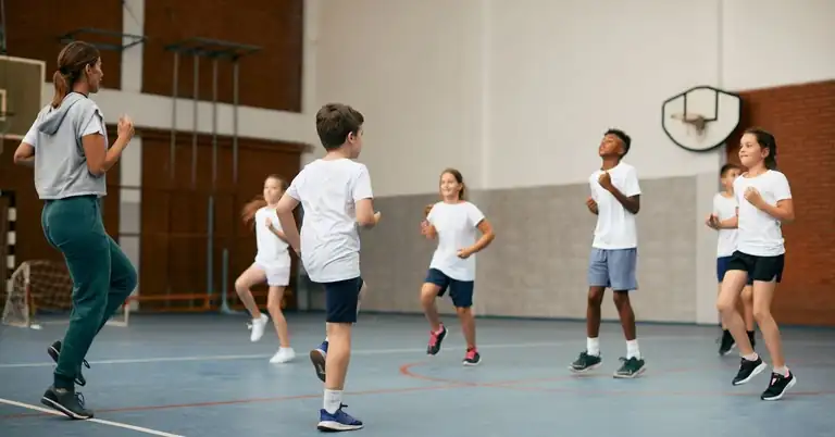 A group of children in a gym are jumping and exercising together. A coach in athletic wear observes and guides them. The gym has a brick wall