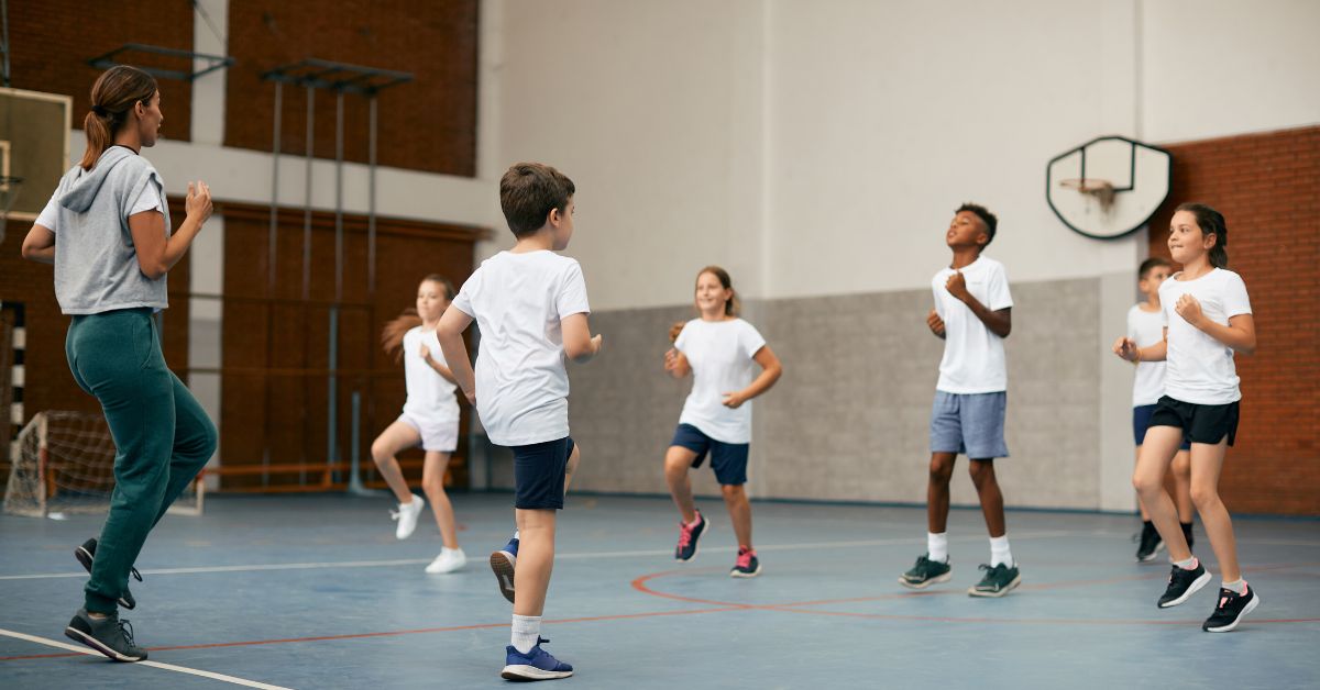 A group of children in a gym are jumping and exercising together. A coach in athletic wear observes and guides them. The gym has a brick wall