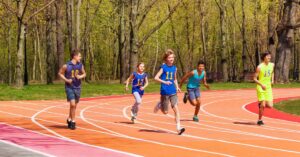 Long Term Athletic Development|Five young athletes running on an outdoor track surrounded by trees. They are wearing various blue and neon outfits