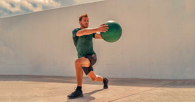Medicine Ball Training|Manfred Scholich Medicine Ball Training|Medicine Ball Books|Medicine Ball Mid Torso Rotation|Assess2Perform Application|Medicine Ball Exercises|Medicine Ball Exercises|A man in athletic wear performs a medicine ball exercise in a gym with a red and black wall. He is mid-action
