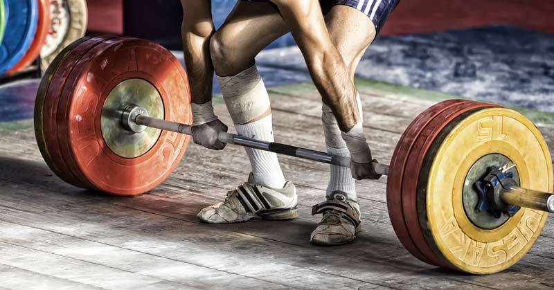 Olympic Lift|Back Development|EMG Snatch|A person in a bright shirt is preparing to lift weights in a gym. They stand over a barbell loaded with colorful plates on a platform. The gym is equipped with racks