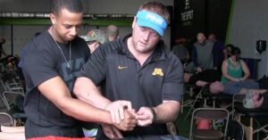 Reflexive Performance Reset|Derrick Suss Pulls Hamstring in Relay|John Fox Ends Sprinting Career with Hamstring Injury|A large group of people wearing black shirts stands in a semi-circle inside a gym. They appear to be listening to a speaker. Most shirts display the word TRAIN in bold letters. Exercise equipment is visible in the background.