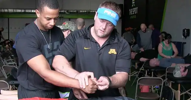 Reflexive Performance Reset|Derrick Suss Pulls Hamstring in Relay|John Fox Ends Sprinting Career with Hamstring Injury|A large group of people wearing black shirts stands in a semi-circle inside a gym. They appear to be listening to a speaker. Most shirts display the word TRAIN in bold letters. Exercise equipment is visible in the background.