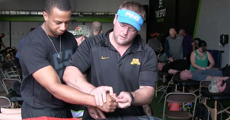 Reflexive Performance Reset|Derrick Suss Pulls Hamstring in Relay|John Fox Ends Sprinting Career with Hamstring Injury|A large group of people wearing black shirts stands in a semi-circle inside a gym. They appear to be listening to a speaker. Most shirts display the word TRAIN in bold letters. Exercise equipment is visible in the background.