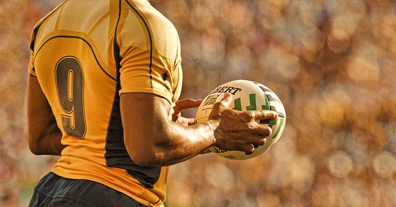Rugby Player Holding Ball|Rugby Players with Ball|A man in athletic gear stands on an indoor track