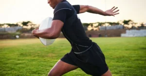 A person in a black athletic outfit runs on a grassy field while holding a rugby ball. The sun sets in the background