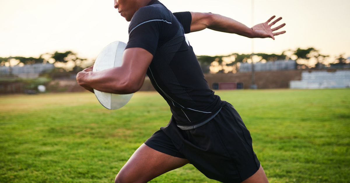 A person in a black athletic outfit runs on a grassy field while holding a rugby ball. The sun sets in the background