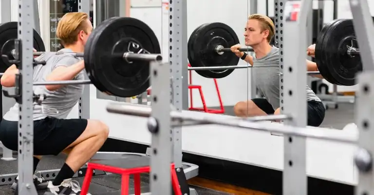 Person doing a squat with a barbell in front of a mirror at a gym. They are wearing a gray shirt and black shorts. The focus is on their form as they look determinedly into the mirror.|Three images: 1) Group of young men in a gym preparing for weightlifting. 2) A person lifting weights with a coachs assistance. 3) An athlete performing a sprint start on a football field.|A person is performing a barbell squat in a gym