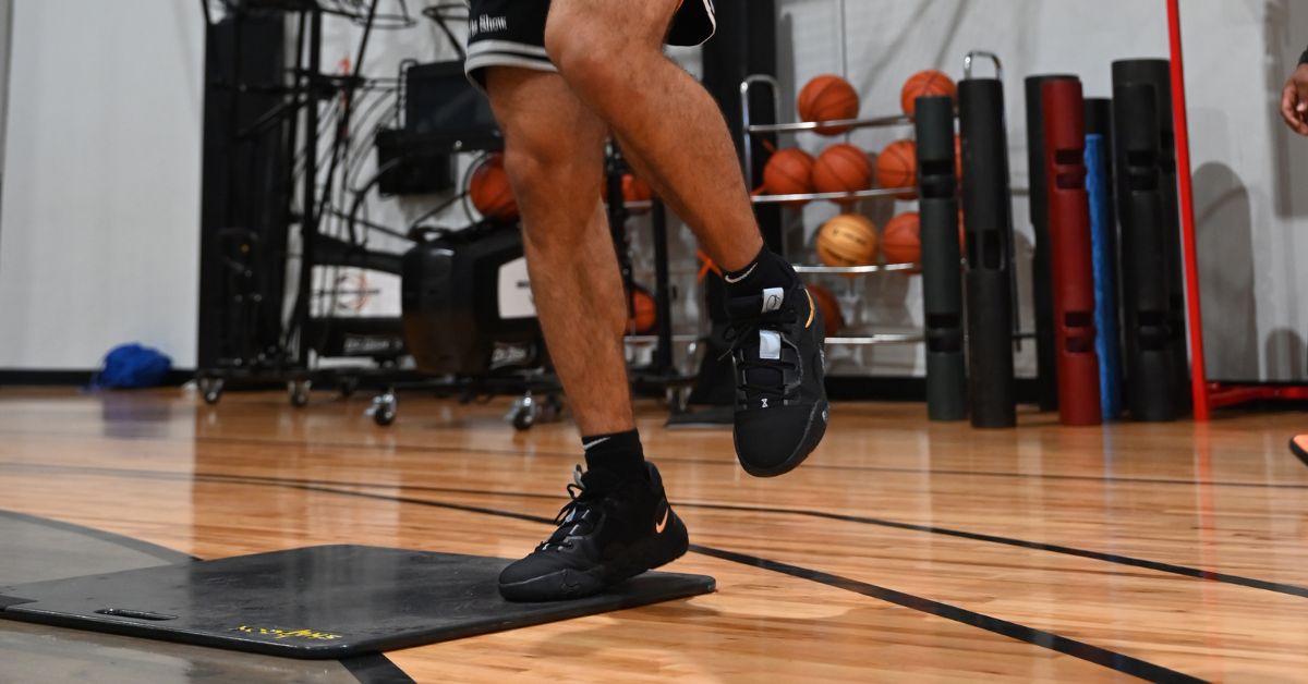 Skyhook Jump Training|A person in a black shirt and jeans holding a large black carrying case with a handle. The case has the word skyhook on it in yellow text. The background features a brick wall and dark carpet flooring.