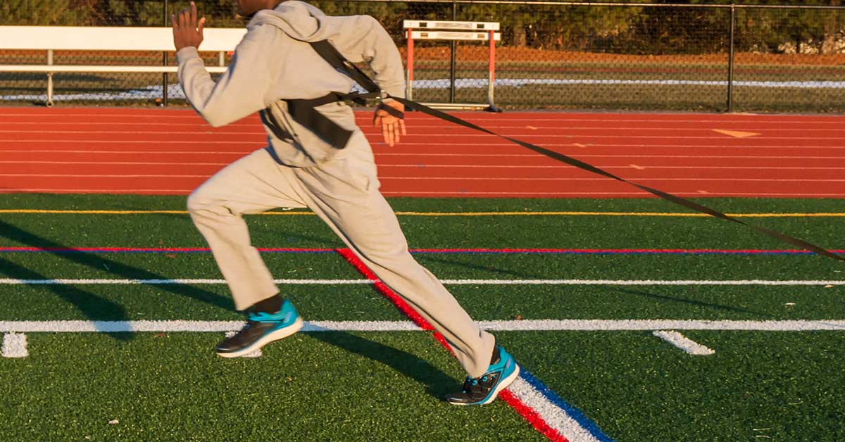 A person in a gray tracksuit sprints on a track while wearing a resistance harness. The track is outdoor