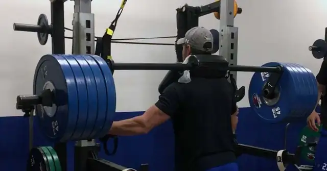 Split Squats|CoachMePlus Heat Map|HSRFESS Strength Phases 1 through 3|HSRFESS Strength Phases 4 and 5|Seasonal RFE Chart|HSRFESS|A man in a black shirt and blue shorts is preparing to lift a heavily loaded barbell on a squat rack. Two other people are nearby