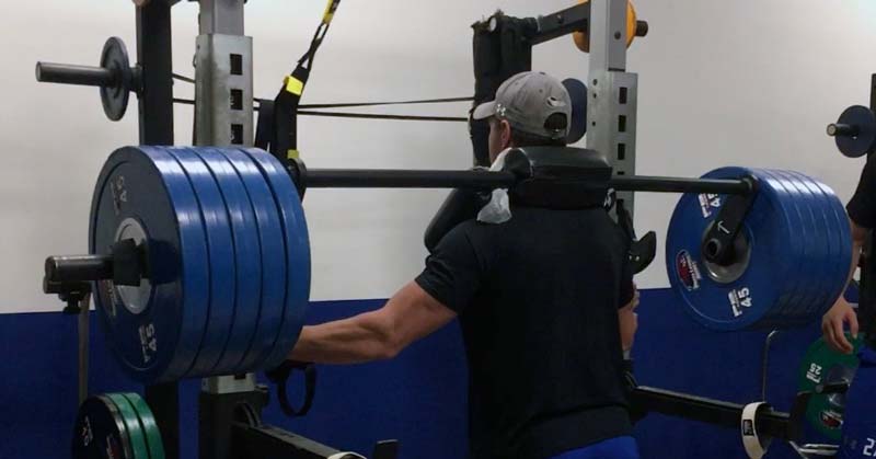 Split Squats|CoachMePlus Heat Map|HSRFESS Strength Phases 1 through 3|HSRFESS Strength Phases 4 and 5|Seasonal RFE Chart|HSRFESS|A man in a black shirt and blue shorts is preparing to lift a heavily loaded barbell on a squat rack. Two other people are nearby