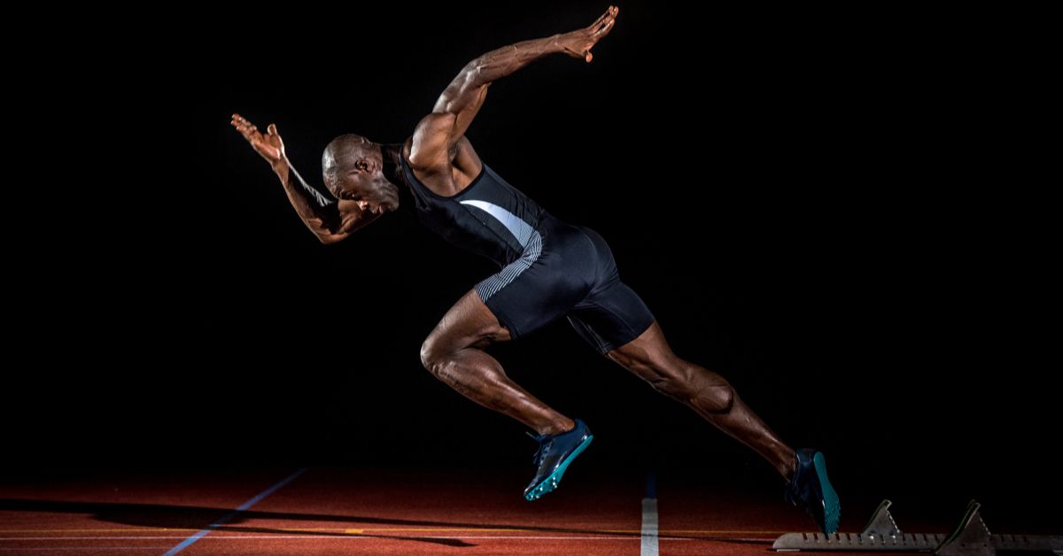 Sprint Start|A person on a red running track is preparing starting blocks. They are outside on a cloudy day with trees and a road in the background.|A person in athletic attire