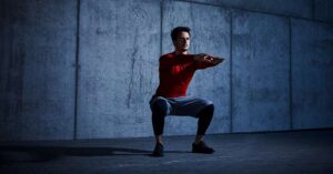 Squat from the ground up|Tall Tree and Squating|A person in a red jacket and black shorts sits on a green turf inside a gym. Dumbbells are lined up in the background