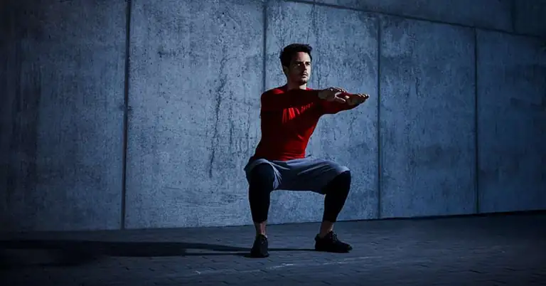 Squat from the ground up|Tall Tree and Squating|A person in a red jacket and black shorts sits on a green turf inside a gym. Dumbbells are lined up in the background