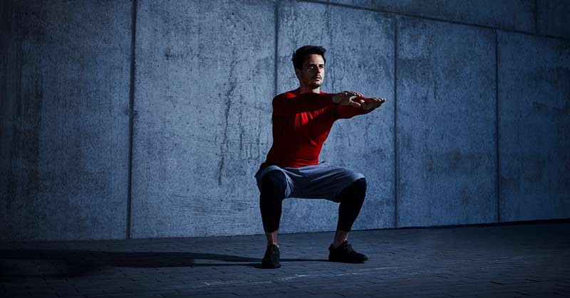 Squat from the ground up|Tall Tree and Squating|A person in a red jacket and black shorts sits on a green turf inside a gym. Dumbbells are lined up in the background