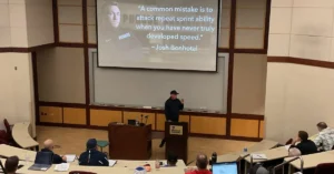 TFC Tony Holler Speed|Tradition and Disruption|Brad Dixon Presentation|A man presents slides to a seated audience in a classroom. The top slide is titled Tier Functional Movement Patterns