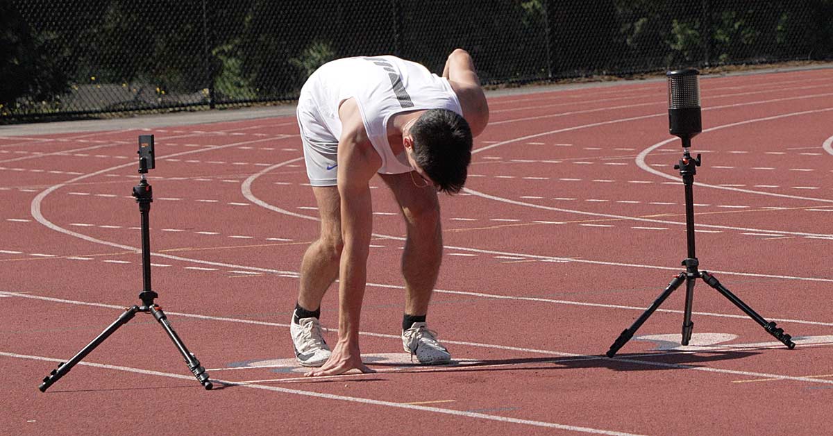 VALD Smart Speed|A male athlete in a white tank top and shorts is sprinting on a track. Two timing devices are positioned in front of him. The background features green turf and trees.|A person in athletic gear prepares to sprint at the starting line on a track. Another person in a black shirt stands nearby