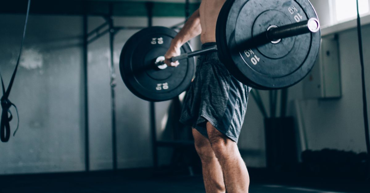 Velocity-Based Training|Close-up of a gym barbell loaded with red weight plates. The background shows a workout area with stacked weight plates