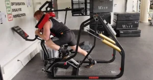Man using a specialized exercise machine in a gym. He is in a leaning forward position with padded supports. Equipment and exercise mats are visible in the background.|Two fitness machines: one for enhancing jump height with a platform and harness