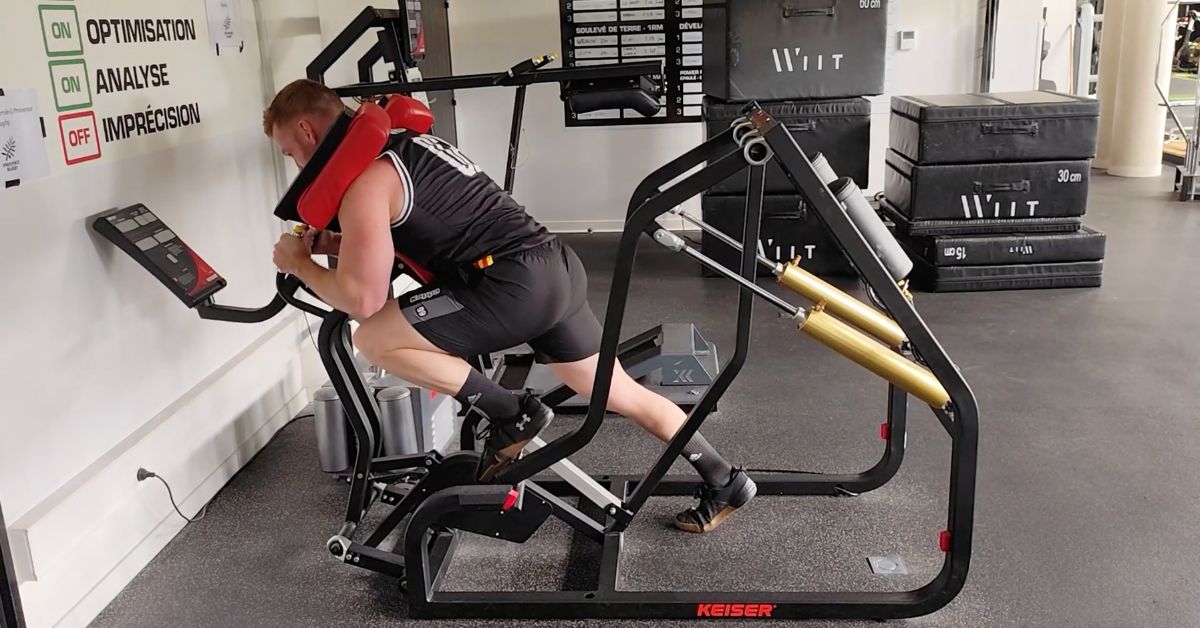 Man using a specialized exercise machine in a gym. He is in a leaning forward position with padded supports. Equipment and exercise mats are visible in the background.|Two fitness machines: one for enhancing jump height with a platform and harness