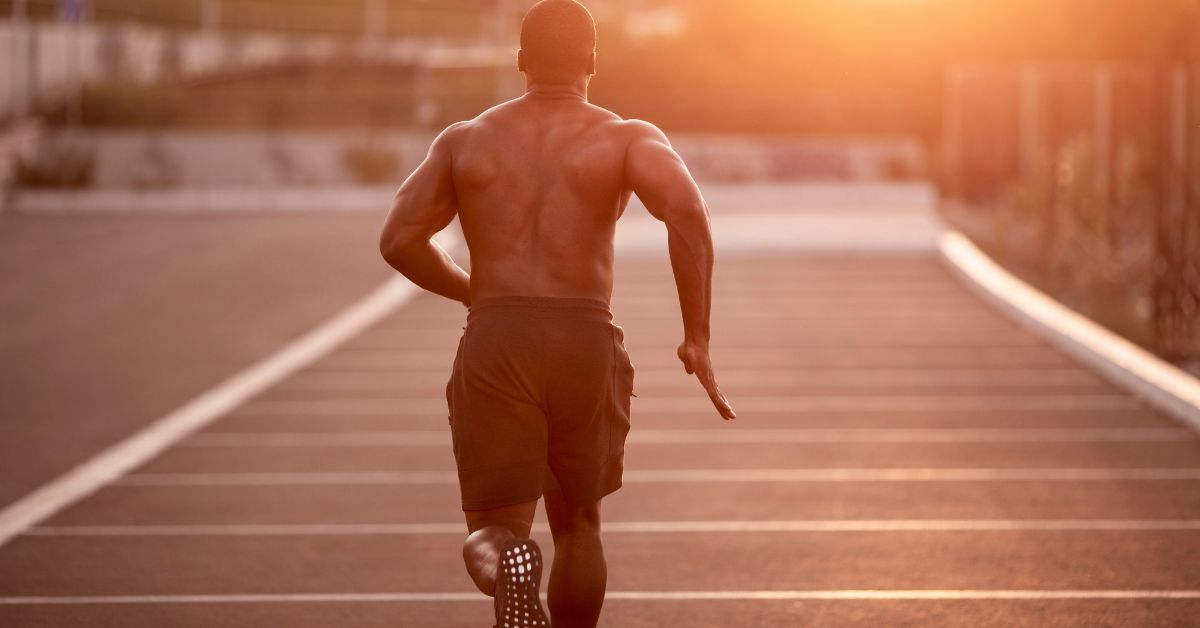 A shirtless man runs on an empty road at sunset. The sunlight creates a warm glow