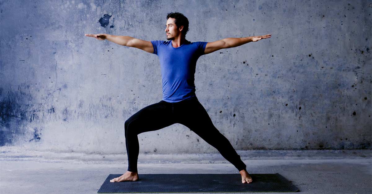Yoga Warrior Pose|A person in a blue shirt and black pants performs a yoga warrior pose on a black mat. The background is a rough concrete wall