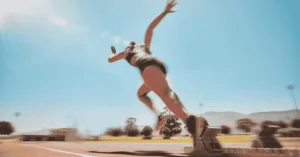 A sprinter is captured in motion, launching from starting blocks on an outdoor track under a clear blue sky, with mountains, trees, and buildings visible in the background.