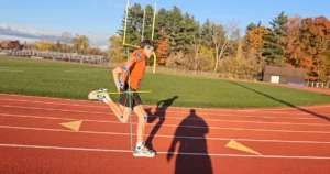 A runner in an orange shirt and black shorts is sprinting on a track. Blue dots and lines mark their joints to analyze running form and adaptive speed. Trees and a field are in the background under a blue sky.