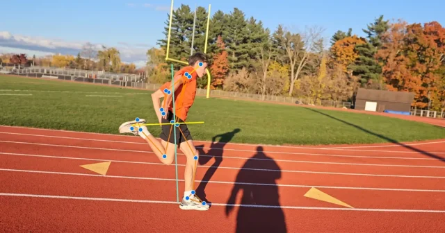 A runner in an orange shirt and black shorts is sprinting on a track. Blue dots and lines mark their joints to analyze running form and adaptive speed. Trees and a field are in the background under a blue sky.